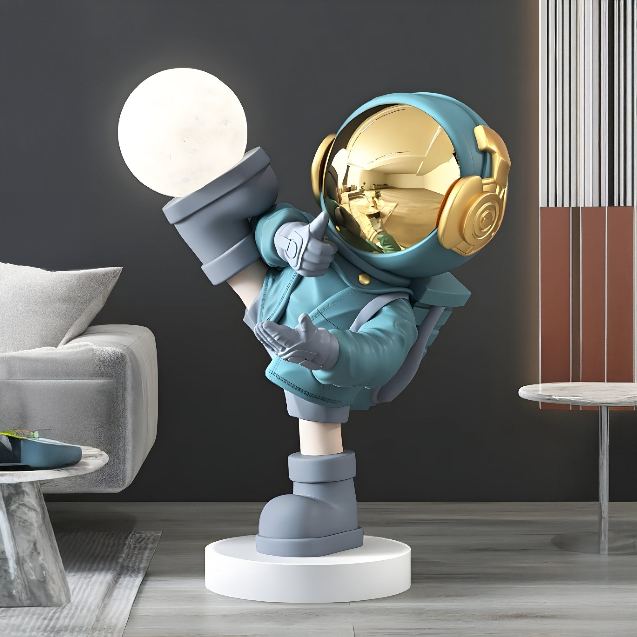 The 70cm Galactic Blue Astronaut Glow Kick Sculpture by Giant Sculptures features a playful design with a shiny gold helmet and glowing orb, balancing on one leg on a white circular platform in a modern room with adjustable lighting, near a gray sofa and striped wall.