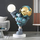 The 70cm Galactic Blue Astronaut Glow Kick Sculpture by Giant Sculptures features a playful design with a shiny gold helmet and glowing orb, balancing on one leg on a white circular platform in a modern room with adjustable lighting, near a gray sofa and striped wall.