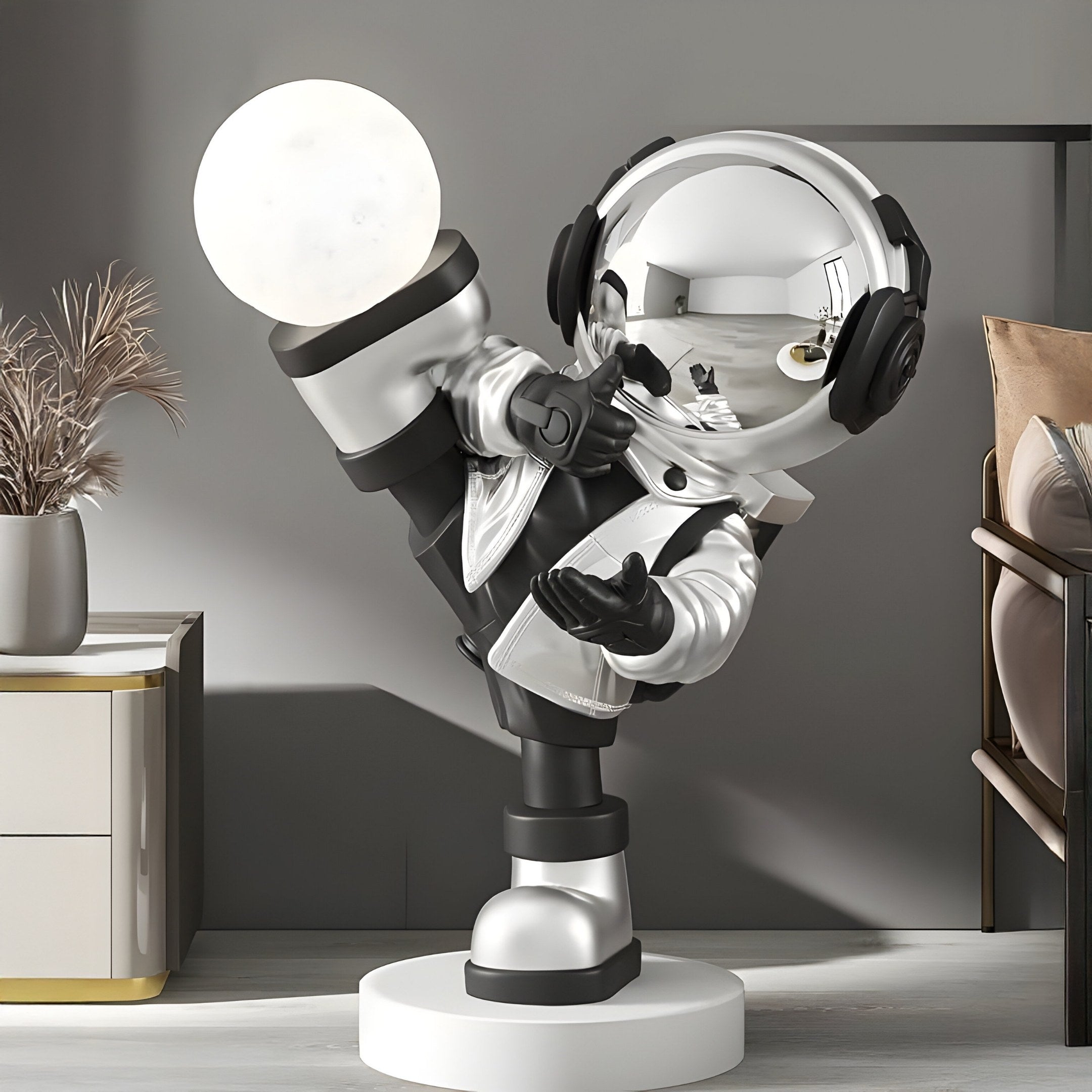 The Giant Sculptures Lunar Silver Astronaut Glow Kick Sculpture, 70cm, showcases a playful pose with an astronaut balancing a lit sphere on its foot, adjustable lighting colors, in a modern setting with neutral tones, alongside a plant, cabinet, and cushioned chair.