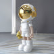 The Cosmic White Astronaut Buddy Sculpture by Giant Sculptures is a 70cm figure with a sleek futuristic look, featuring a gold helmet visor and white spacesuit. It stands indoors holding a gold object while wearing large white boots.