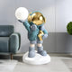 A 75cm Galactic Blue Astronaut from Giant Sculptures, on a white base cradling a Glow Orb Sculpture, features a reflective helmet, teal suit, gray boots, and holds a gold star. A gray sofa and green potted plant complete the space-themed décor.