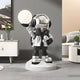 The Giant Sculptures Lunar Silver Astronaut Beats Glow Orb Sculpture - 75cm, with a shiny helmet and headphones, adorns a modern living room. It rests on a round base, enhancing the neutral tones and contemporary furniture with its captivating space-themed accent.