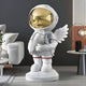 The Nova White Astronaut Angel Rocket Sculpture by Giant Sculptures stands 75cm tall, featuring a silver astronaut with wings, a golden helmet visor, and holding a rocket. Its the perfect cosmic addition beside a gray sofa and round table with a spherical lamp in any modern living room.