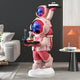 The Stellar Pink Astronaut Duo Server Sculpture by Giant Sculptures (78cm) serves drinks on a modern tray in a stylish living room, placed beside a gray couch and an orange table lamp.