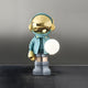 The Galactic Blue Astronaut Glow Orb Sculpture by Giant Sculptures is an 80cm figure with a shiny gold helmet and glow orb. It wears a teal jacket, gray shorts, and boots, standing on a textured surface under ambient lighting against a gray backdrop.