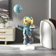 In a modern room lit by ambient lighting, the Galactic Blue Astronaut Butler Balloon Sculpture - 80cm by Giant Sculptures features a golden-helmeted butler balancing a tray and polka-dotted balloon, standing gracefully on a white base with a cabinet and vase of flowers nearby.