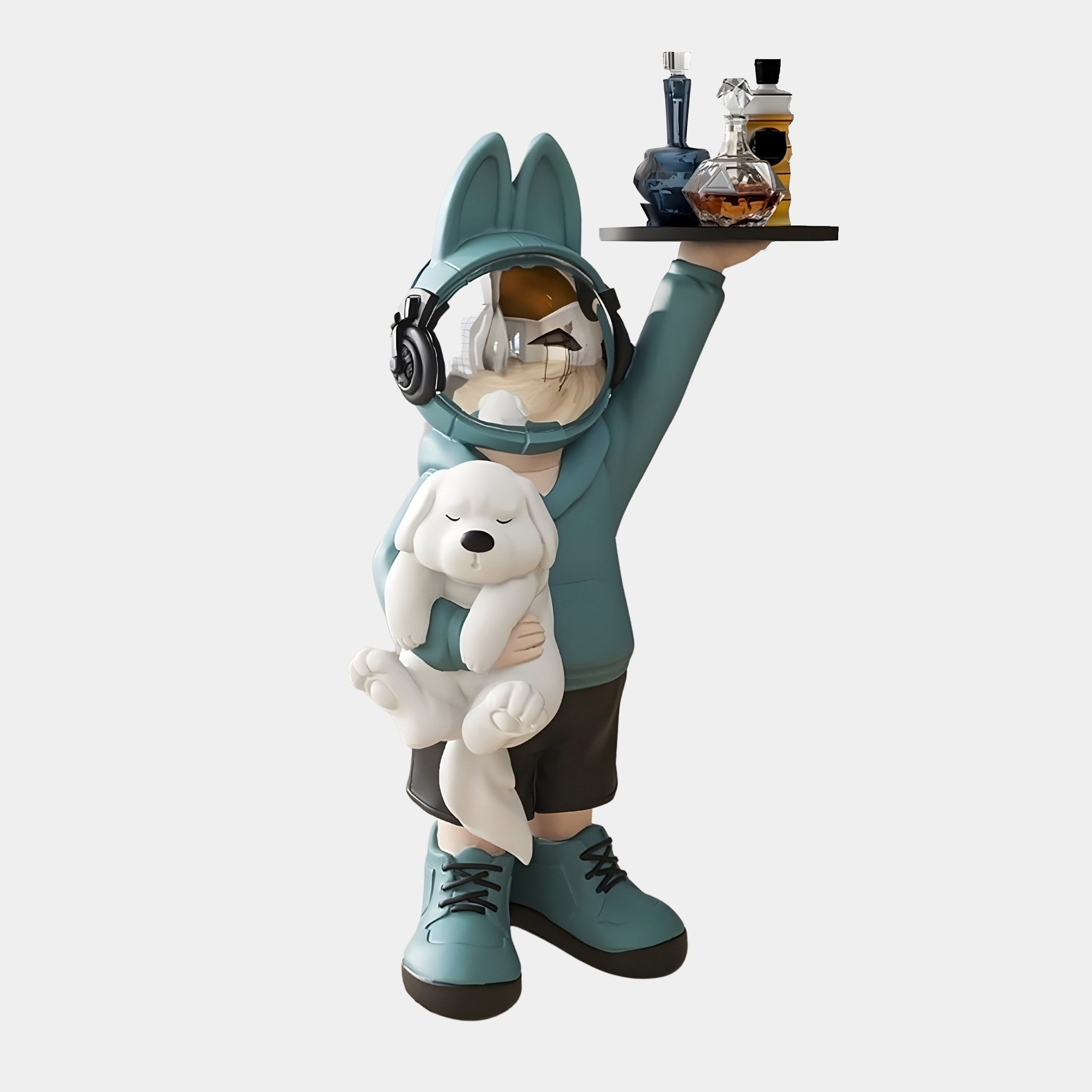 A cartoonish Astronaut Bunny Pup Butler sculpture by Giant Sculptures, standing 80cm tall, features bunny ears and headphones. Its dressed in a blue hoodie, shorts, and shoes while holding a tray of bottles with one hand and cradling a white dog against a plain backdrop.