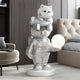 In a modern room, the Chrome Astronaut CosmoPals Glow Bear Sculpture by Giant Sculptures, at 80cm, holds a glowing sphere while a cat sits on its head. The setting is complemented by a beige tufted chair and dark wooden panels.