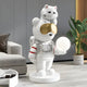 In a modern living room with a sleek sofa, vibrant plant, and elegant wood paneling, the White Astronaut CosmoPals Glow Bear Sculpture by Giant Sculptures adds whimsy as the bear astronaut cradles a glowing sphere while a cat perches on its helmet and the gold-tinted visor glints under soft lighting.