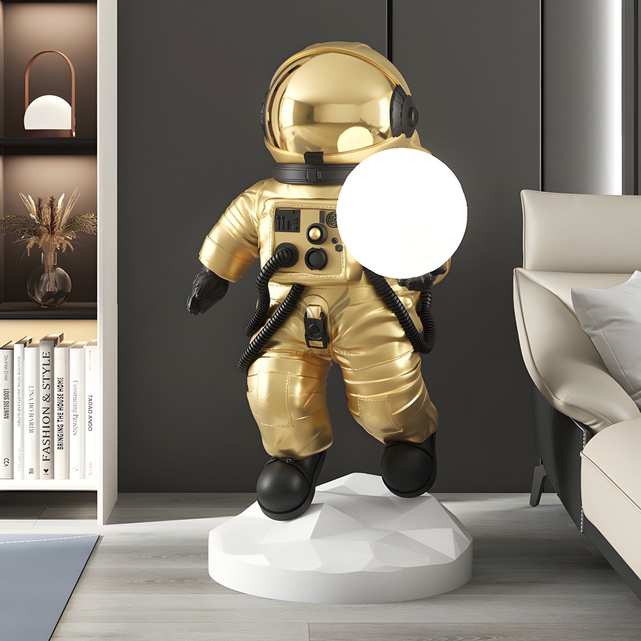 A modern room features the Celestial Gold Astronaut Ranger Glow Orb Sculpture by Giant Sculptures on a white base. On the left is a book-filled shelf, and on the right rests a white cushioned chair. A small brown vase with dried plants enhances this celestial decor.