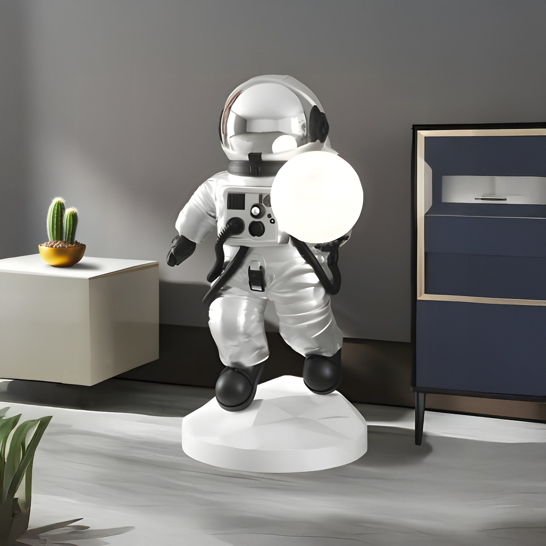 The Giant Sculptures Lunar Silver Astronaut Ranger Glow Orb Sculpture (80cm) stands on a white base, holding a glowing orb. It adds futuristic décor to your space, complemented by a potted cactus on a table and a dark dresser in the background.