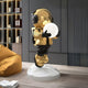 The Celestial Gold Astronaut Glow Hover Sculpture by Giant Sculptures stands on a white platform, holding a glowing orb. Dressed in gold and black with helmet and headphones, it enhances any rooms decor with wooden shelves and a mustard couch, offering adjustable lighting colors to set the mood.