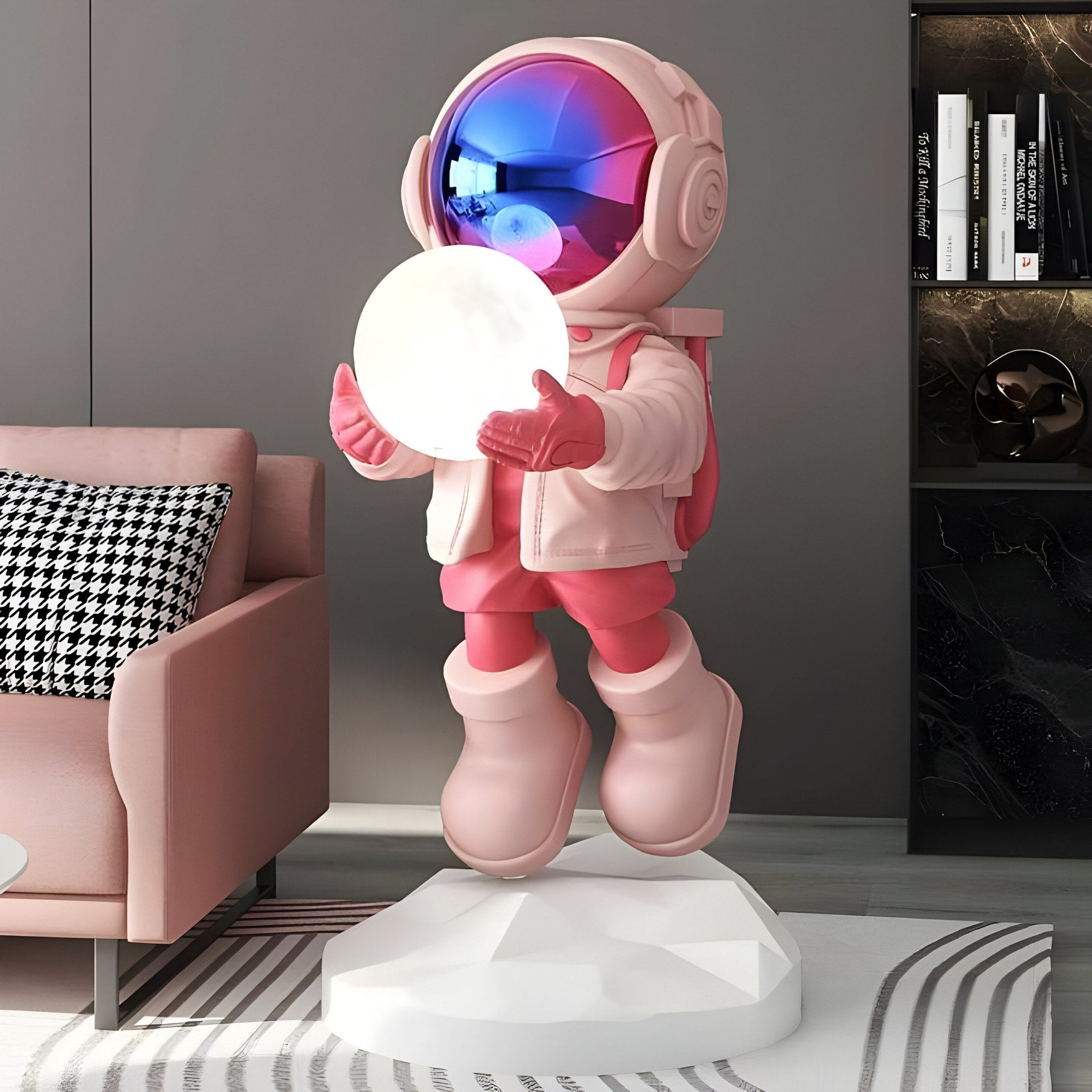 The Stellar Pink Astronaut Glow Hover Sculpture - 80cm by Giant Sculptures, features an adjustable light on a geometric base and holds a glowing orb. Its reflective helmet shines in blue and red, set against a modern living room with books, a pink couch, and a checkered pillow.