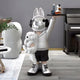 The Silver Astronaut Bunny Pup Butler Sculpture by Giant Sculptures, standing 80cm tall, features a silver helmet with rabbit ears. It holds a white dog and a tray with bottles, wearing a gray hoodie and black shorts, perfect to complement modern decor with an avant-garde flair.