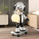 The 80cm Lunar Silver Skater Astronaut Glow Sculpture by Giant Sculptures shows a headphone-wearing astronaut with a glowing orb on a skateboard, set indoors amid modern furniture, a potted plant, and a bed.