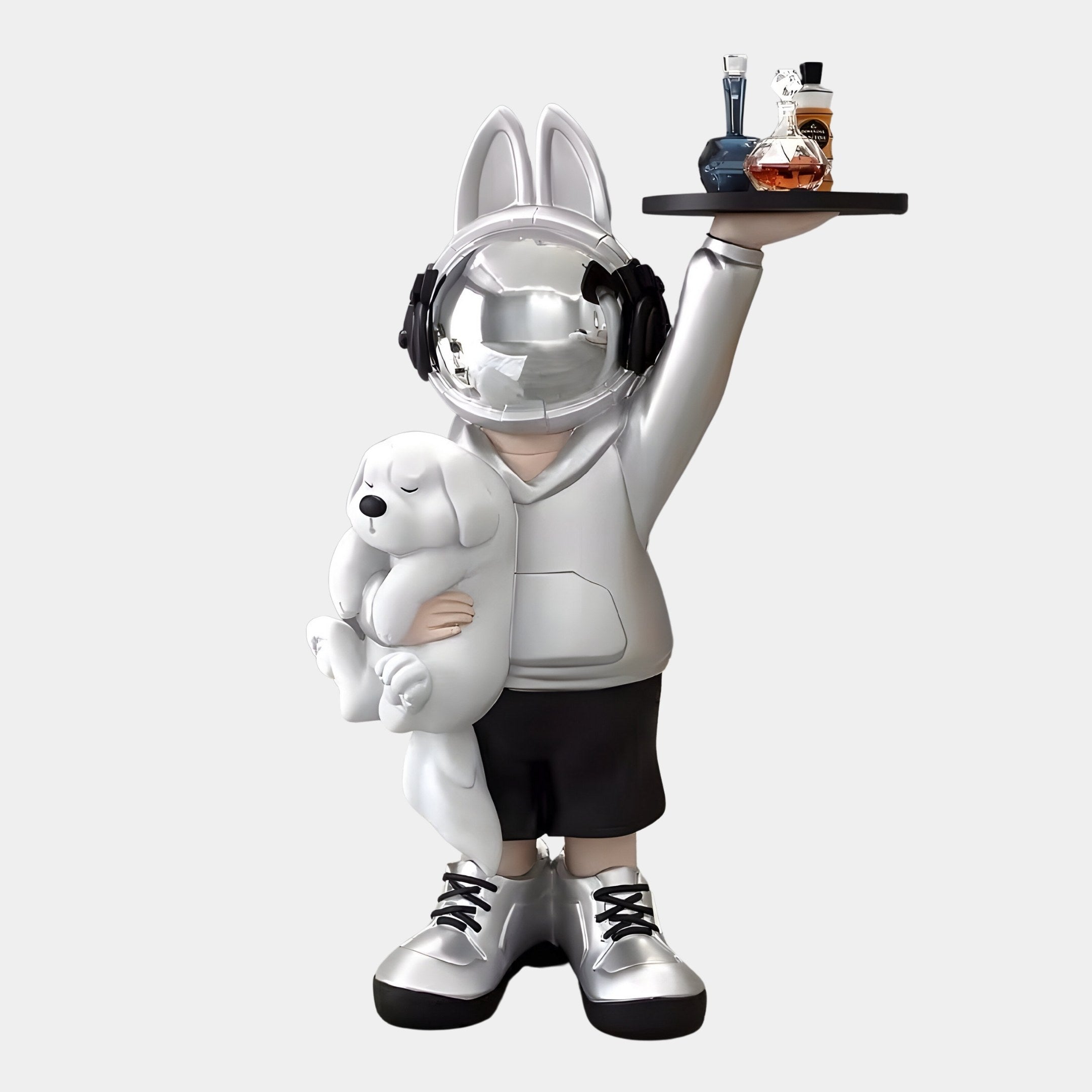 The Silver Astronaut Bunny Pup Butler Sculpture by Giant Sculptures features a child wearing a silver astronaut bunny helmet, headphones, and hoodie while holding a white dog plushie and a tray with bottles. This 80cm piece beautifully blends whimsy with modernity for stunning home decor.