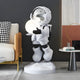 The Nova White Astronaut Glow Hover Sculpture by Giant Sculptures, standing 80cm tall, showcases a futuristic decorative figure holding a glowing orb atop a geometric base. It complements a modern sofa and side table with stacked books, blending dark and light tones in sleek contemporary style.