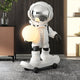 Nova White Skater Astronaut Glow Sculpture by Giant Sculptures, 80cm resin astronaut on a skateboard holding a glowing orb, styled in a modern living room with dark panelled wall and neutral furniture.