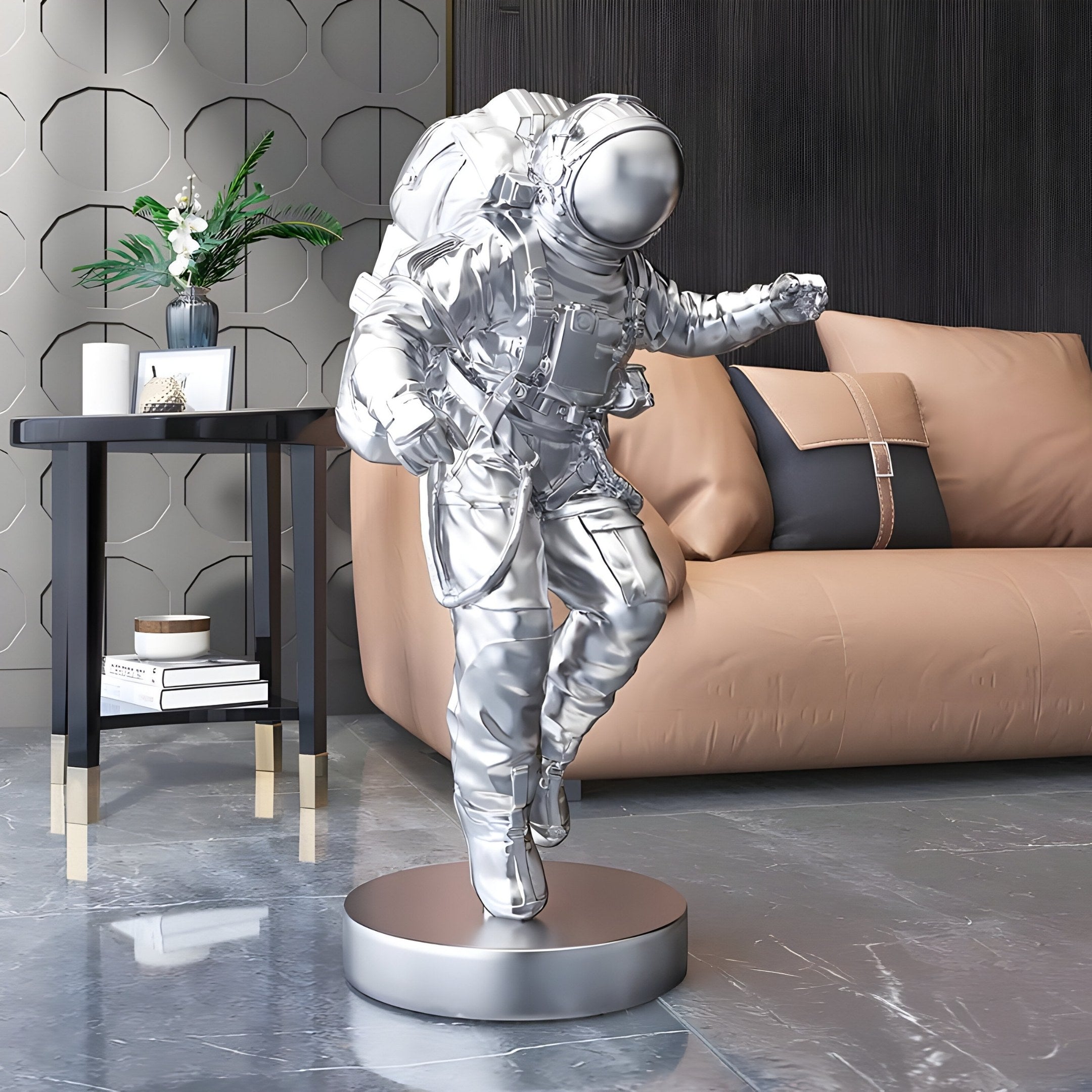 The Cosmic Chrome Space Astronaut Explorer Sculpture by Giant Sculptures stands on a round base in a modern living room. It enhances the space with an imaginative touch, set before a brown couch with pillows, next to a small black table holding a vase and books against a patterned wall.