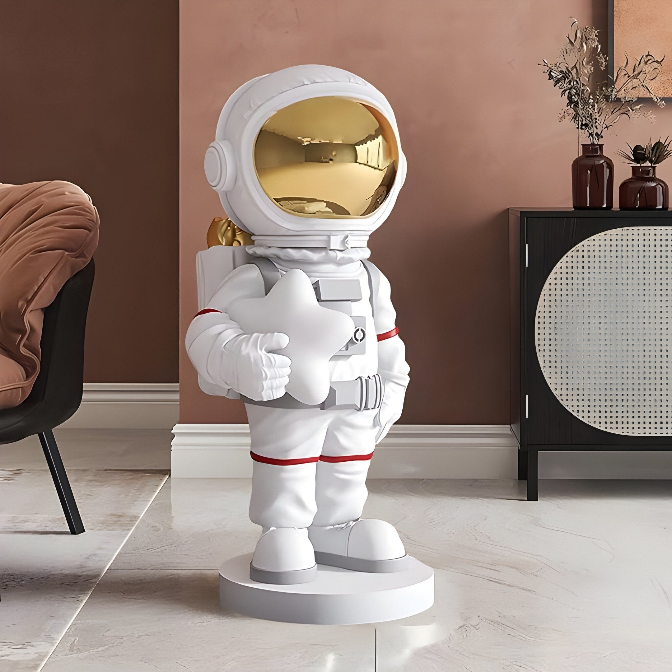 The Nova White Astronaut Star Sculpture by Giant Sculptures, featuring a shiny gold visor, stands indoors on a marble floor. It is surrounded by modern furniture: a black chair and brown cabinet with vases, all set against a warm brown wall for a cosmic décor ambiance. Height: 85cm.