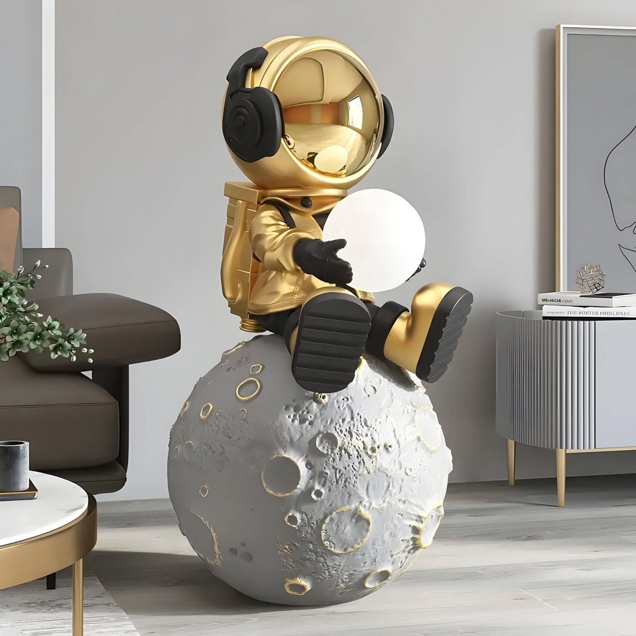 A Celestial Gold Astronaut from Giant Sculptures, wearing a shimmering suit with headphones, sits on the 86cm Glow Moon Sculpture and holds a white orb, set in a modern living room with adjustable lighting, featuring a gray cabinet and abstract art.