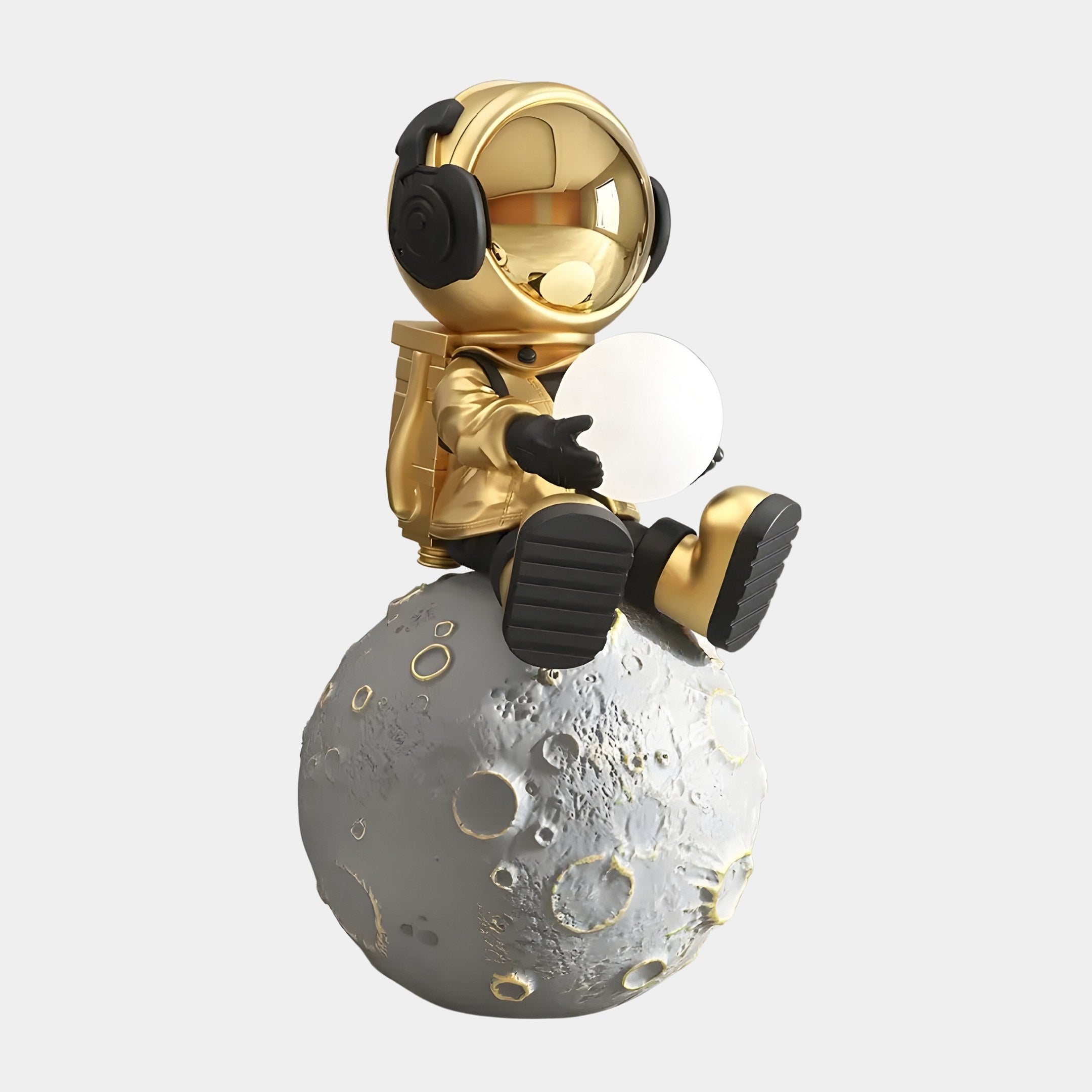 The Celestial Gold Astronaut Glow Moon Sculpture by Giant Sculptures features a golden astronaut with black boots and headphones sitting on a grey, cratered moon, holding an orb. The helmet reflects adjustable lighting for contrast against the textured lunar surface.