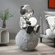 A metallic astronaut figurine with large headphones, clutching a small white disk, sits on the Giant Sculptures 86cm Lunar Silver Astronaut Glow Moon Sculpture in a modern room with minimalistic decor, including a bed and plant.