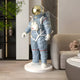 The Celestial Blue Space Astronaut Standing Sculpture (88cm) by Giant Sculptures adds a modern touch to your living room décor—perfect beside sofas and shelves, with plants and décor accents in the background.