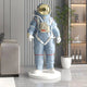 The Giant Sculptures Celestial Blue Space Astronaut Standing Sculpture (88cm) stands life-size on a white base in a modern living room, complementing a gray sofa, tall plant, and shelves with bottles and decor accents.
