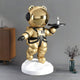 The 90cm Celestial Gold Astronaut Bear Butler Sculpture by Giant Sculptures acts as a futuristic butler on a white platform, tray in paw with a small bottle. It seamlessly integrates into the modern room featuring a wooden cabinet, glass of water, and tall vase with dried flowers.