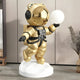 The Celestial Gold Astronaut Bear Glow Sculpture - 90cm by Giant Sculptures features a mirrored helmet, glowing orb, and stands on a white base with backpack and earphones. Set against gray walls and wooden cabinet accents with decorative grasses, it’s perfect for modern décor.