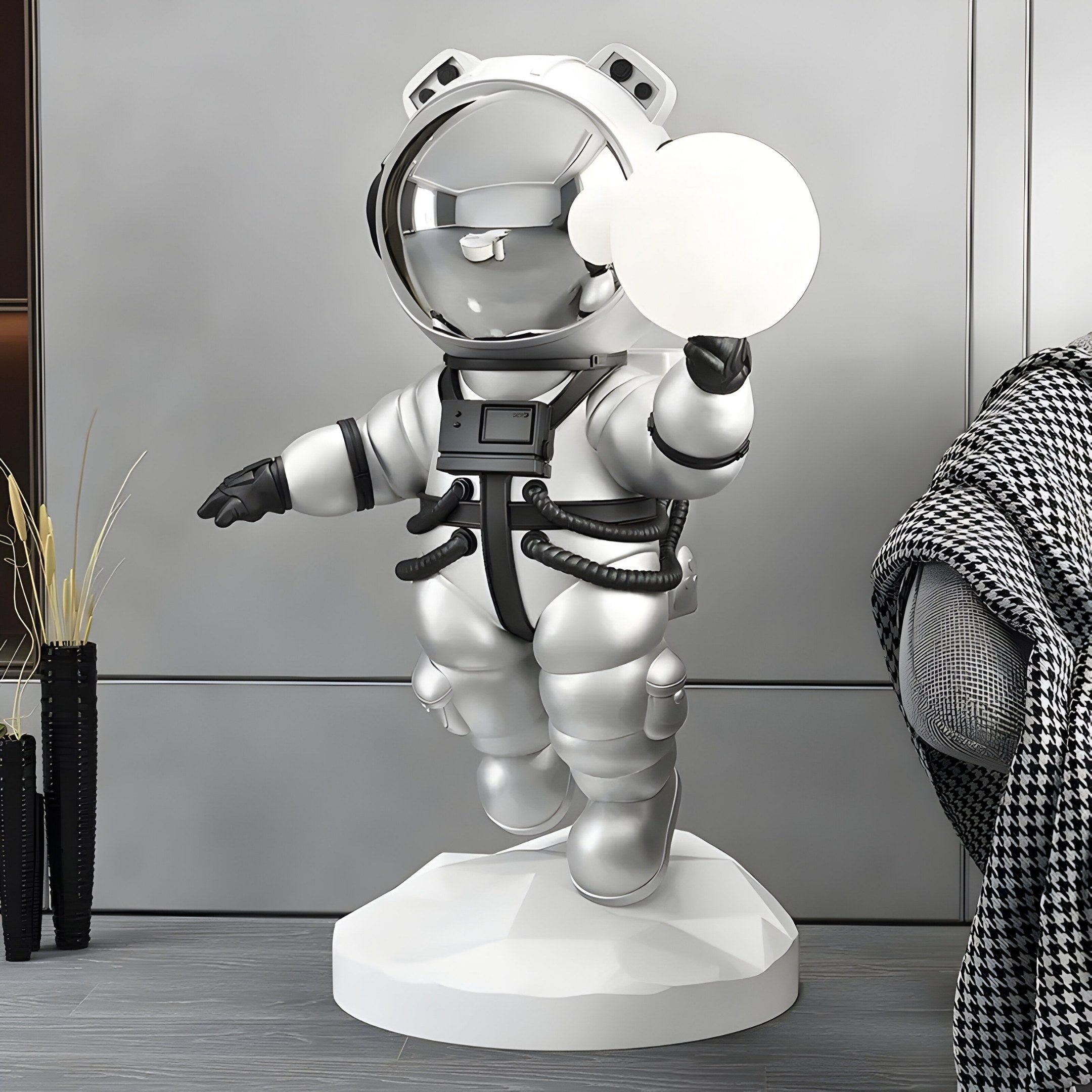 The Astral Silver Astronaut Bear Glow Sculpture by Giant Sculptures, standing 90cm tall, features a silver bear holding a glowing orb on a geometric base. It adds futuristic décor indoors with black accents near plants and a checkered blanket over a gray chair.