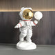 The Nova White Astronaut Bear Glow Sculpture by Giant Sculptures features a 90cm astronaut with a gold visor holding a sphere. The detailed suit, with red accents, stands on a round base against a dark wood background, offering space-themed elements similar to the original design.