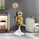 The Celestial Gold Astronaut Rocket Lift-Off Balloon Sculpture by Giant Sculptures stands on a cloud, enhancing a modern living room with its space-themed elegance, perfectly complementing the white couch, checkered pillow, and stylish cabinet.