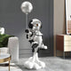 In a modern living room with a sofa, plant, and wooden cabinet, the 95cm Lunar Silver Astronaut Rocket Lift-Off Balloon Sculpture by Giant Sculptures stands on a cloud-like base, its moon-textured design enhancing the space theme ambiance.