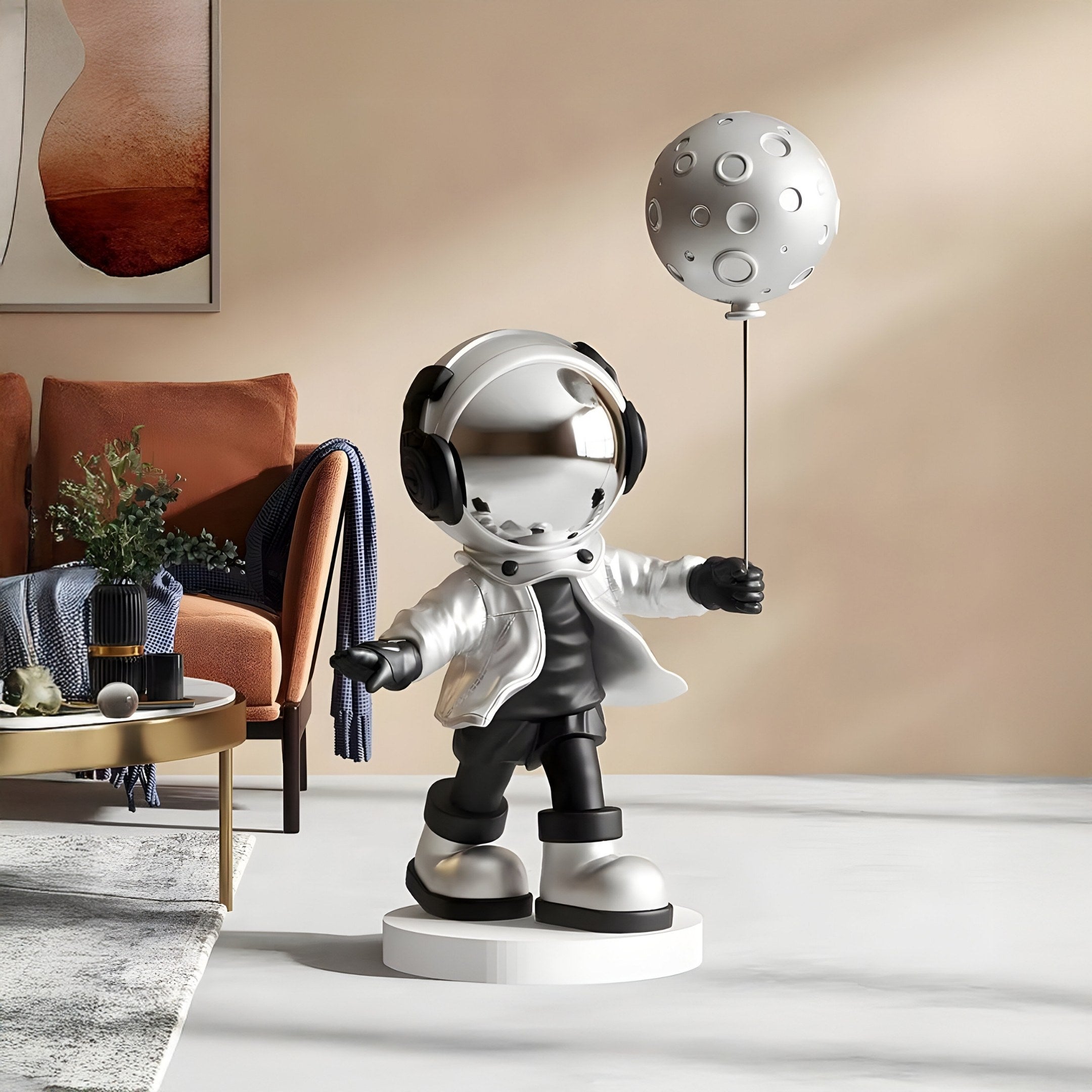 The Giant Sculptures Lunar Silver Astronaut Moon Balloon Sculpture (96cm) stands whimsically on a pedestal with a moon-shaped balloon. Outfitted in a shiny helmet and suit, it complements modern decor featuring a couch, round coffee table, and abstract wall art as an interior centerpiece.