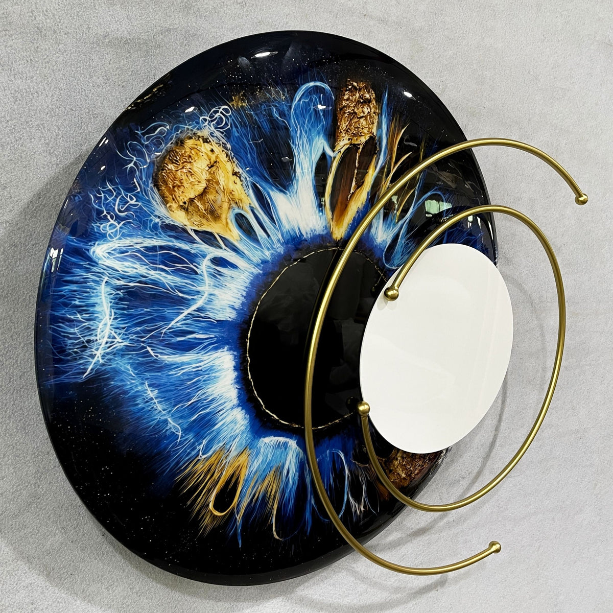 Aurelios Cosmic Blue Crystal Porcelain & Gold Orbit 3D Wall Art featuring a circular blue and brown iris design with a mirrored core and orbiting gold metal rings, shown against a plain grey background.
