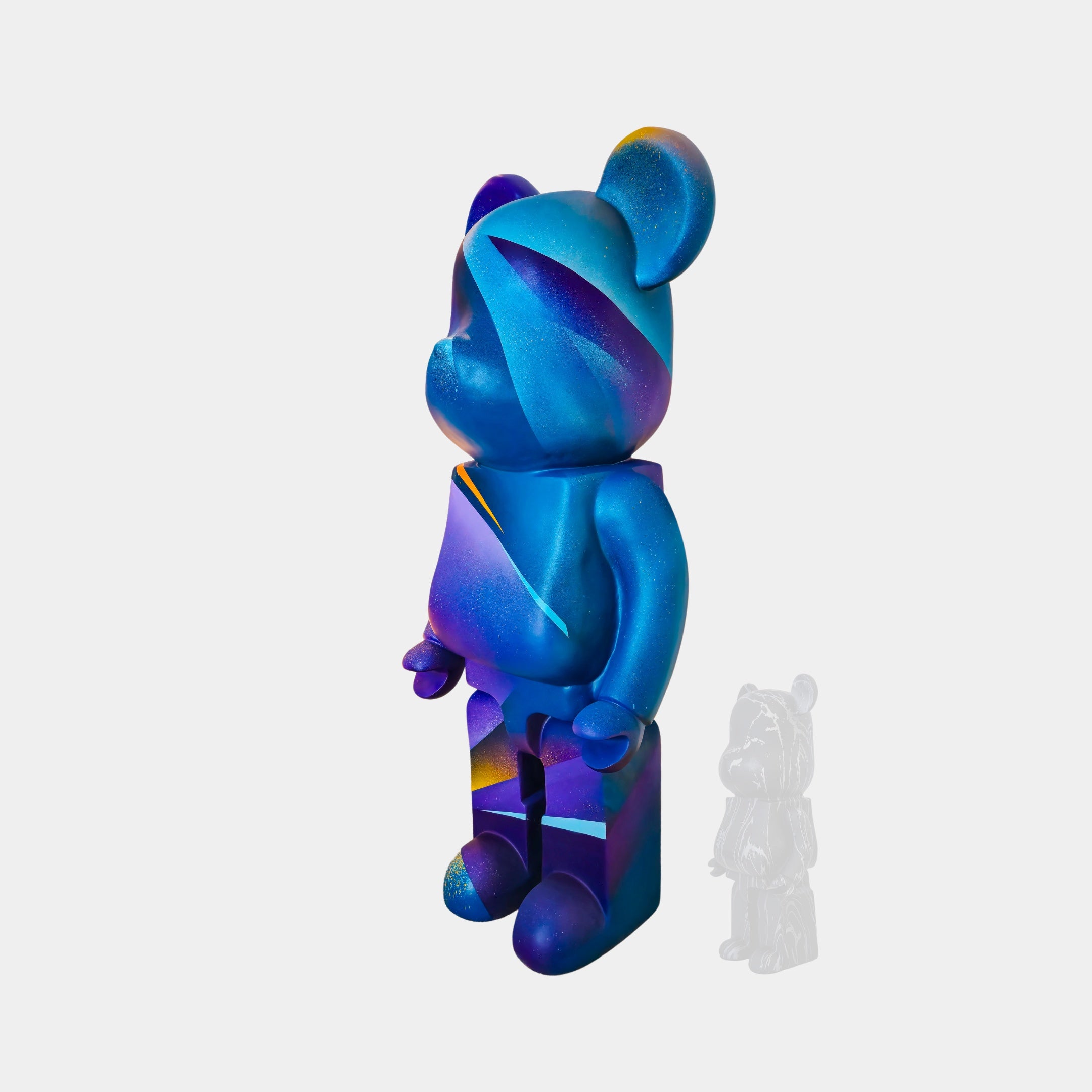 The "Midnight Sunset Bear Sculpture - 165cm" by Giant Sculptures, a blue and purple bear-shaped vinyl figure with abstract patterns, is positioned facing left. Behind it, against a white background, stands a smaller, semi-transparent gray version of the bear, highlighting its status as an exclusive art collectible by Adam Illes.