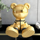 The Celestial Gold Astronaut Bear Sculpture by Giant Sculptures, clad in a futuristic outfit and visor, sits on a black shelf. Plant leaves creep into the frame on the left, while partially visible magazines with Asian characters on the right add to this imaginative décor.