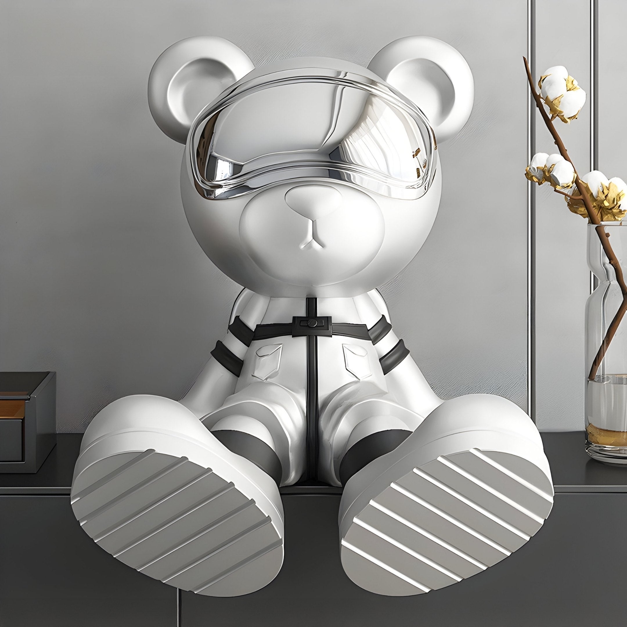 The 51cm Cosmic Chrome Astronaut Bear Sculpture by Giant Sculptures, featuring a reflective visor and harness accessory, rests against a gray wall. Its glossy white finish is complemented by a vase with cotton branches on the right, adding an organic touch to the scene.