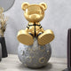 The Celestial Gold Astronaut Bear Moon Sculpture by Giant Sculptures features an astronaut bear in a spacesuit on a textured gray moon. This space décor is accented with decorative plants and a circular wall mirror, arranged artistically on a light-colored surface.