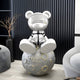 The 80cm Cosmic Chrome Astronaut Bear Moon Sculpture by Giant Sculptures features a futuristic bear in a helmet and suit on a moon-like sphere, framed by a black ridged wall with a minimalist shelf, bare branch, and touches of Moon Base against the light floor and chair.