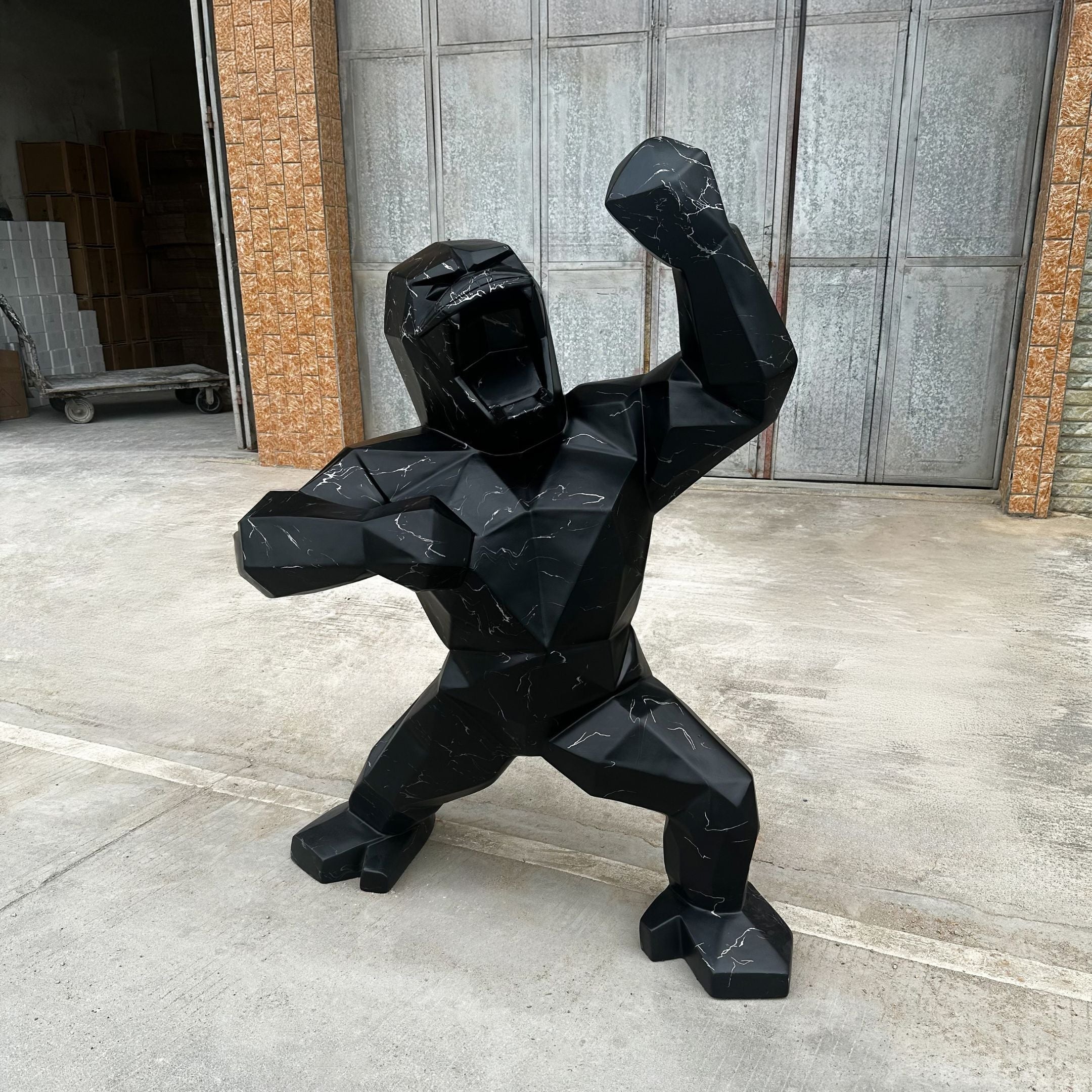 The Black Stone Gorilla Sculpture - 165cm by Giant Sculptures is placed outdoors on a concrete surface in front of a metallic industrial-style door. Its large, geometric form with raised arms and an open mouth showcases modern design, suggesting a roaring pose.