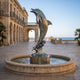 Life-Size Contemporary Leaping Dolphin Bronze Sculpture (250cm) with warm patina, arched mid-leap above a coral-form base, set in a stone fountain on a coastal building terrace.