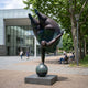 Giant Contemporary Voluptuous Acrobat on Sphere Bronze Sculpture, 250cm, dark bronze figure balanced on hands atop a sphere with legs split overhead, displayed outdoors on a stone base near a glass building and trees.