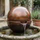 Contemporary Hammered Sphere Ball Bronze Fountain, 60cm, dark brown patina sphere with flowing water in a stone basin, set in a courtyard garden with lavender and potted plants.
