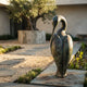Monumental Modern Crane Bird Bronze Sculpture by Giant Sculptures with arched neck and green-brown patina, set on stone paving in a courtyard with olive tree.