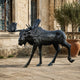 Monumental Walking Moose Bronze Sculpture by Giant Sculptures shown in mid-stride with raised foreleg and wide antlers, dark patina, set on a stone courtyard.