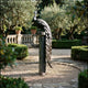 Large Traditional Peacock Bronze Sculpture by Giant Sculptures on a tall stone column, tail cascading down, set in a formal garden with olive trees.