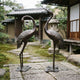 Large Traditional Crane Pair Bronze Sculpture by Giant Sculptures, dark patinated cranes on lotus bases in a Japanese garden with tiled gate and stone lantern.